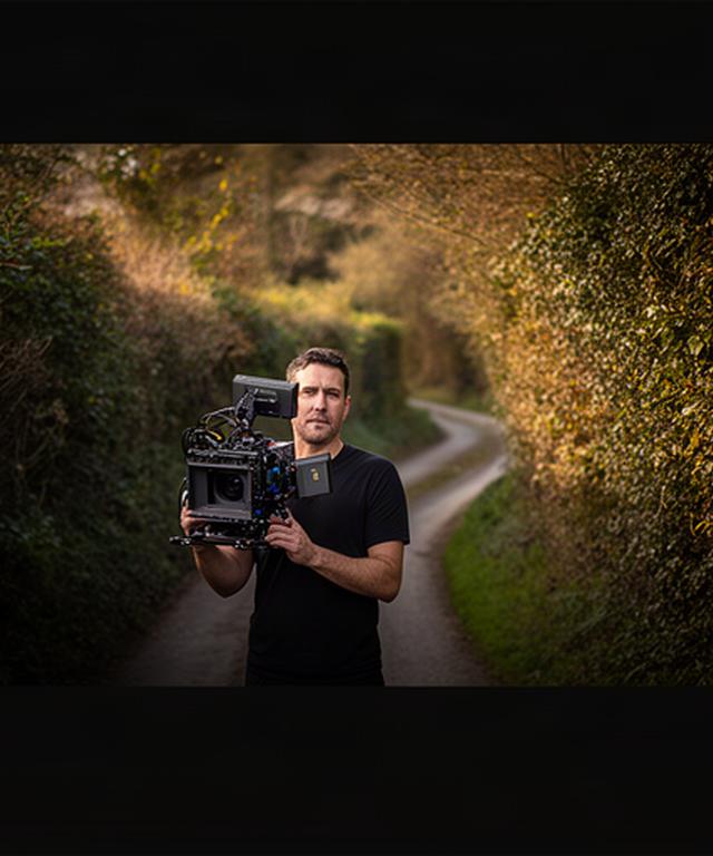 Video producer holding a cinema camera beside a Devon hedgerow on Peek Lane