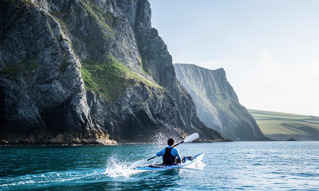 Kayaker moving through calm blue water beneath rugged Devon cliffs on an outdoor campaign shoot
